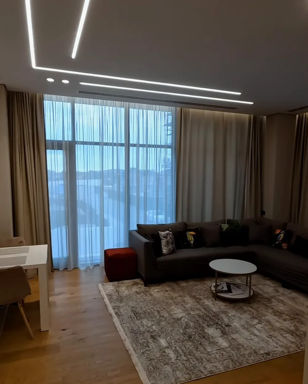 The living room of an apartment in Lighthouse: a corner sofa, floor-to-ceiling windows, light lines on the ceiling
